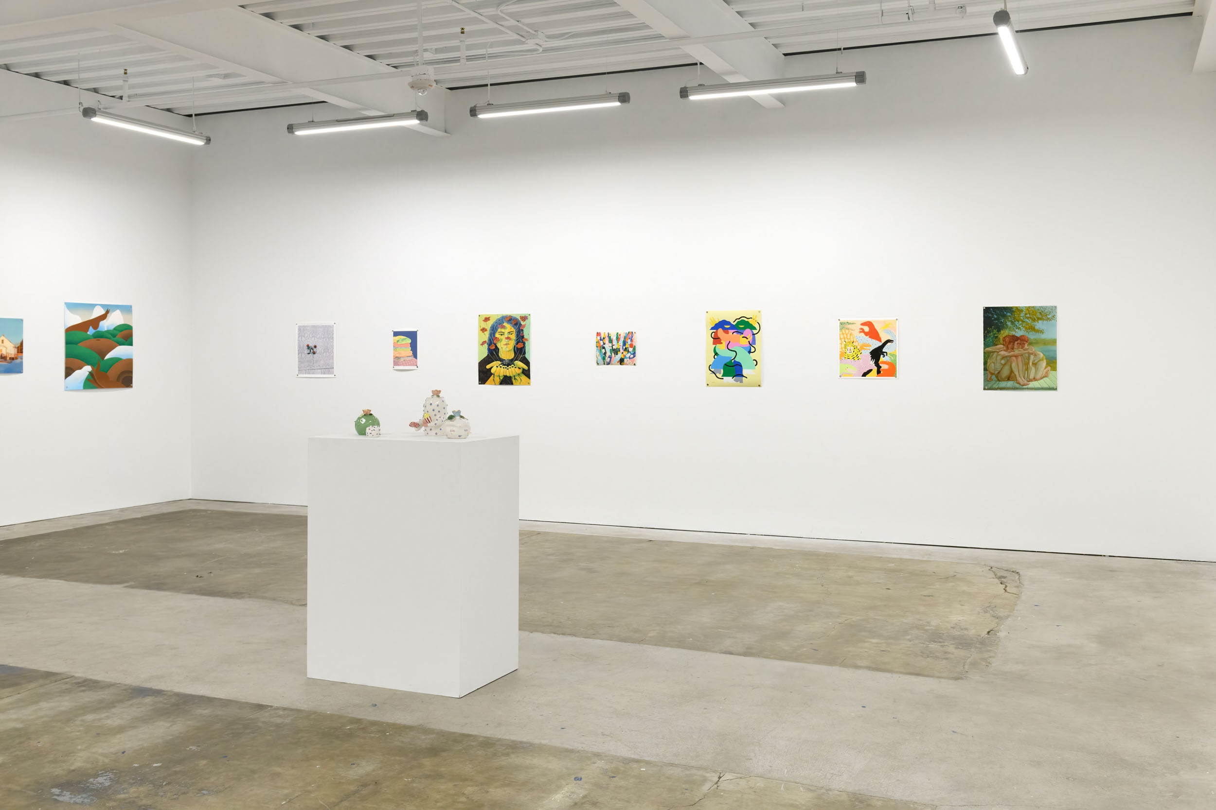 Prints of artworks on a white wall in an art gallery with fluorescent lighting. 